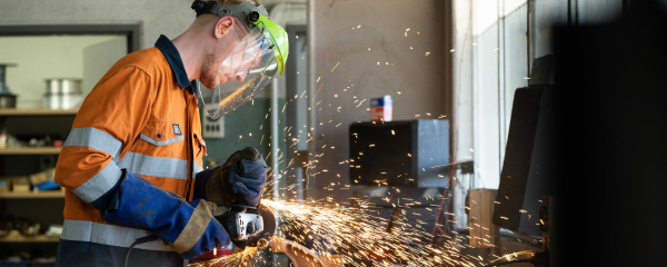Man wearing an orange hi-vis and face shield using an angle grinder, sending out sparks, in a workshop